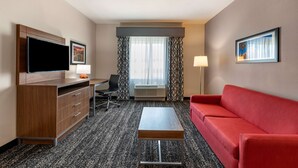 Ein Hotelzimmer mit Flachbildfernseher, einem Holzkonsolenmöbel, einem roten Sofa, einem kleinen Tisch, einem Schreibtisch mit Stuhl und einem großen Fenster mit gemusterten Vorhängen.