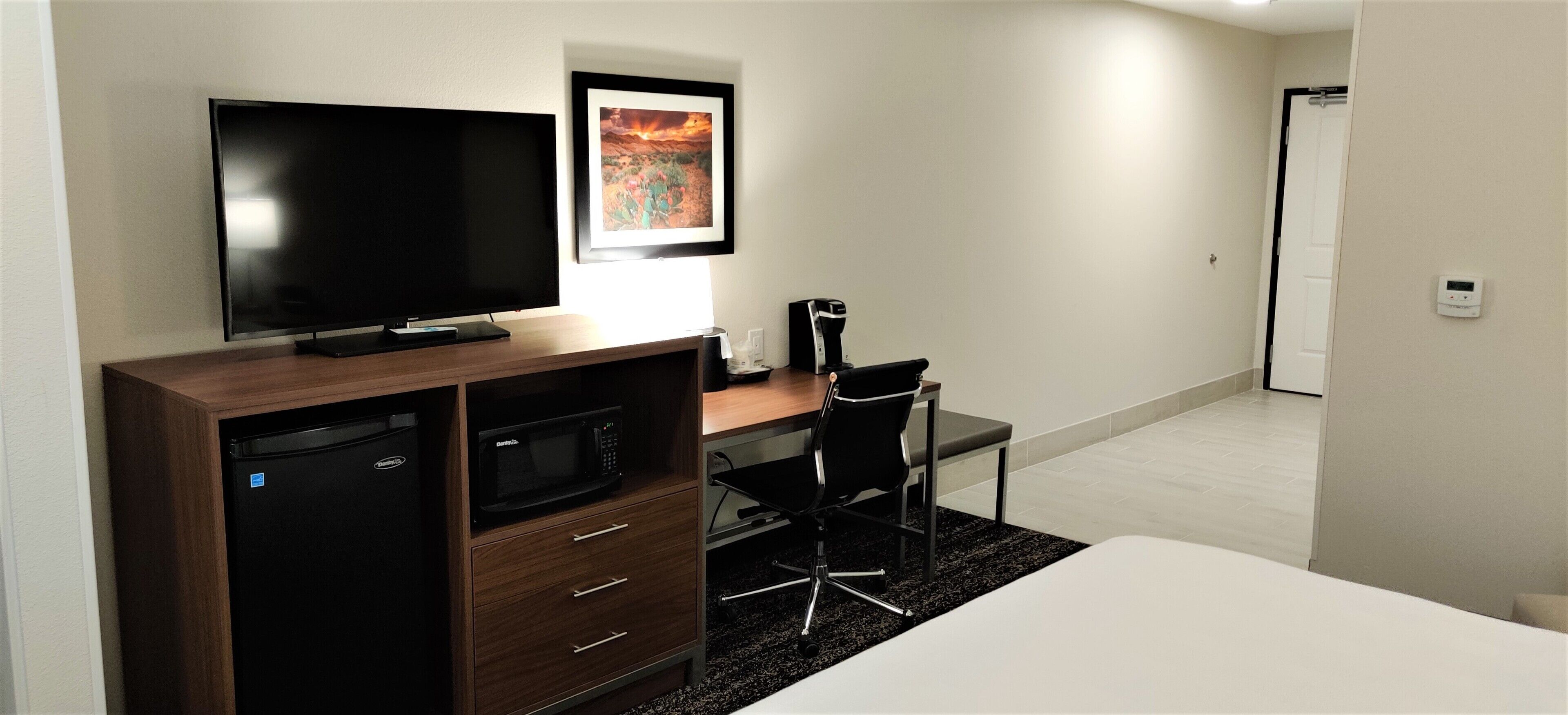 A modern hotel room with a flat-screen TV, a microwave, a coffee maker, a desk with a chair, and a bed.