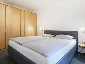 A double bed with white linens, a wooden wardrobe, and a bedside table with a lamp.