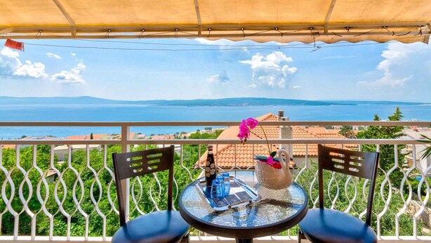 Un balcon avec une table et des chaises, offrant une vue panoramique sur la mer et les montagnes lointaines.