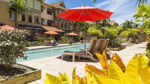 Un espace piscine doté de chaises longues et de parasols, entouré d’une végétation luxuriante et bordé d’un immeuble à plusieurs étages avec des balcons.