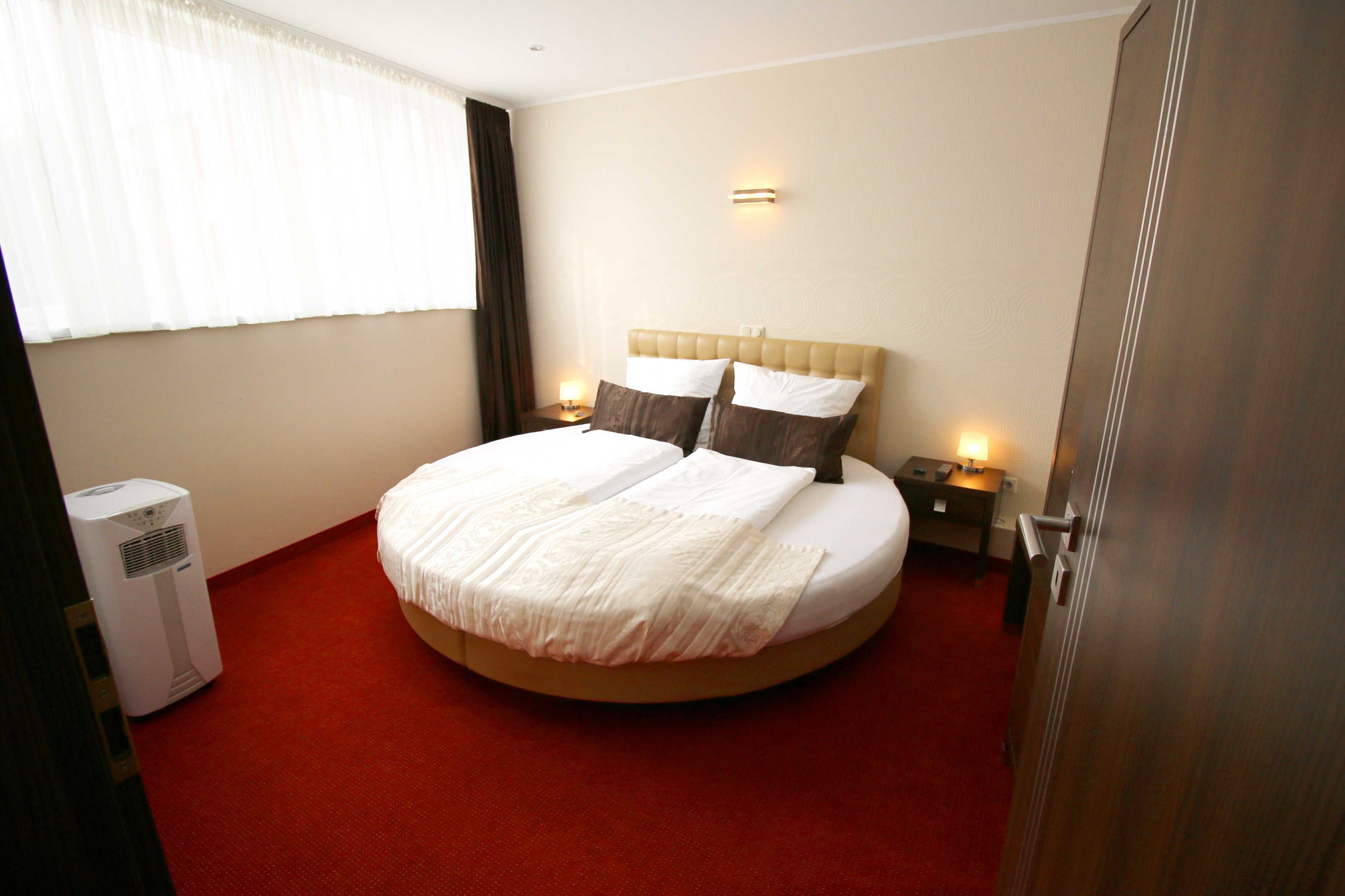 A hotel room with a round bed, two bedside tables with lamps, a wall-mounted light, and a window with curtains.
