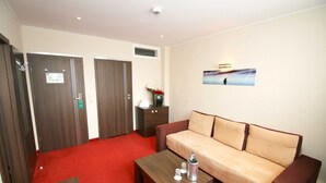 A hotel room with a red carpet, a beige sofa, a wooden coffee table, and two wooden doors. - Junior suite with bath - Home Hotel (Wilhelmshaven)