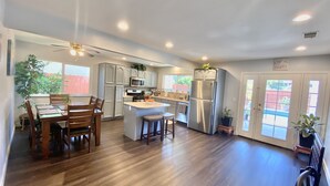 A modern kitchen with a central island, wooden dining table, and chairs. There are cabinets, a refrigerator, and a ceiling fan. - Kids Love Us! Legoland Home with A Pool (Oceanside)