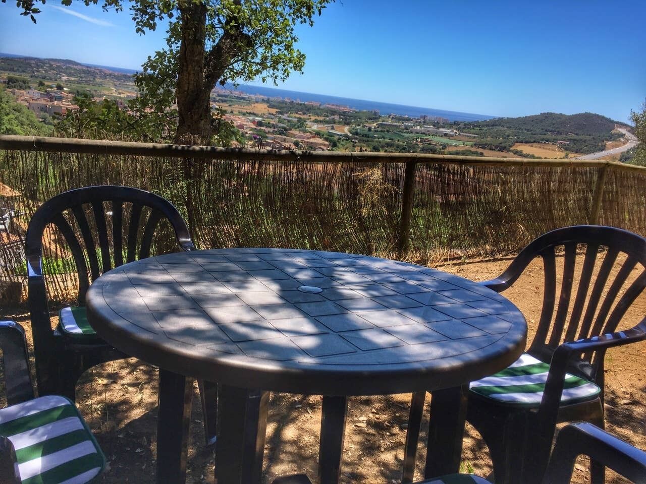 A round table with chairs in a shaded outdoor area, overlooking a scenic landscape with hills and a distant view of the sea.