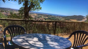 Mesa redonda com cadeiras em área externa sombreada, com vista para uma paisagem cênica com colinas e o mar ao longe.