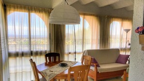 Uma sala de jantar com uma mesa, cadeiras e um sofá. Há uma janela grande com cortinas, um lustre e vista para o exterior.