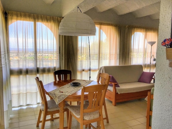 A dining area with a table, chairs, and a sofa. There is a large window with curtains, a hanging light fixture, and a view of the outdoors.