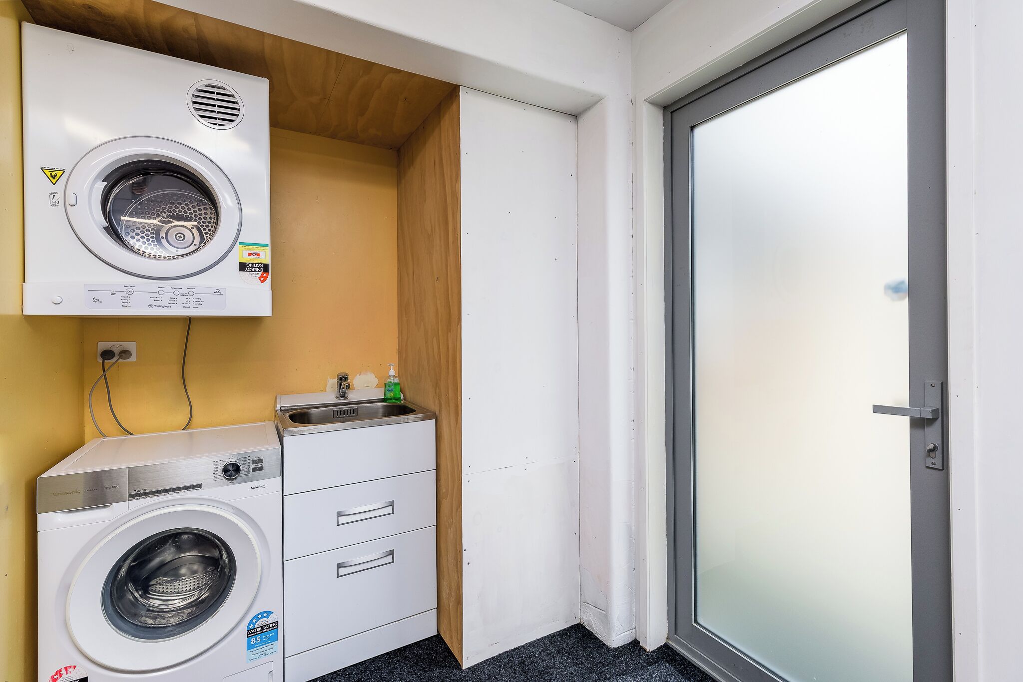 A compact laundry room with a front-loading washing machine and a dryer, a small sink with a soap dispenser, and a door with a frosted glass panel.