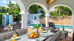 Un espacio exterior para comer con mobiliario de mimbre, piscina y un muro de piedra.