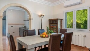 Un comedor con una mesa dispuesta para cuatro personas, rodeada de sillas, un frutero y un aparador decorativo. Un arco de entrada conduce a una cocina con salpicadero de azulejos y una ventana con vista a follaje.