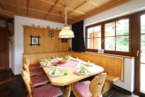 A wooden dining room with a large table set for four, wooden chairs, a hanging light fixture, and a window with a view of buildings and greenery.