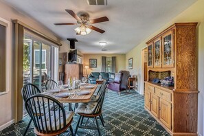 A dining area with a wooden table set for six, surrounded by chairs. There is a ceiling fan, a fireplace, and a cabinet with glass doors.