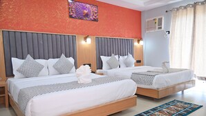 A hotel room with two beds, each with a grey headboard, white bedding, and patterned pillows. There is a blue patterned rug on the floor, a wall-mounted air conditioner, and a decorative wall piece above the beds.