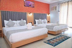 A hotel room with two beds, each with a grey headboard, white bedding, and patterned pillows. There is a blue patterned rug on the floor, a wall-mounted air conditioner, and a decorative wall piece above the beds.