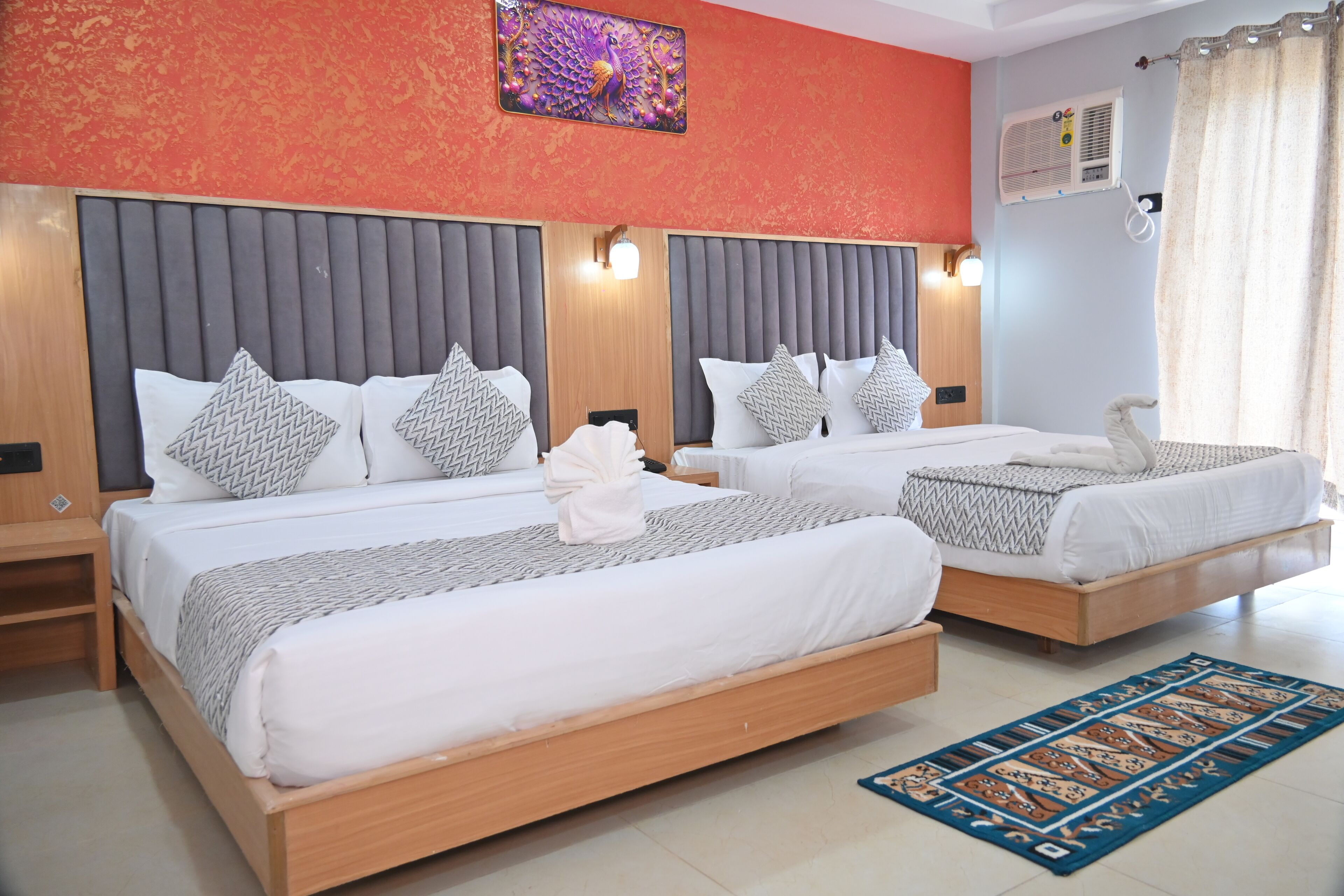 A hotel room with two beds, each with a grey headboard, white bedding, and patterned pillows. There is a decorative wall piece above the beds and a blue patterned rug on the floor.