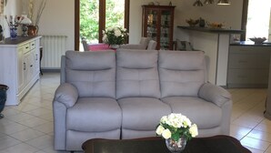 Una sala de estar con un sofá gris, una mesa de centro de madera con un jarrón de flores, y una cocina al fondo.