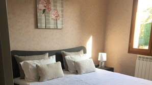 Una cama bien hecha con una colcha blanca y varias almohadas, una lámpara de noche y una obra de arte en la pared.