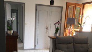 Una sala de estar con un sofá gris, una mesa de centro de madera y una lámpara. Se ve una puerta al fondo que conduce a otra habitación.