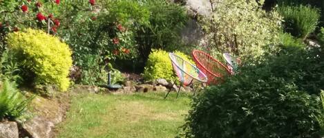 A garden with various plants and a stone wall. There are colorful chairs and a small table.
