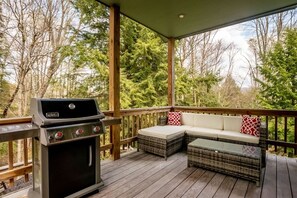 Eine überdachte Terrasse im Freien mit Grill, bequemen Sitzgelegenheiten und Blick auf Bäume.