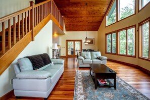 A spacious living room with a wooden staircase, two sofas, a coffee table, and large windows offering a view of the outdoors.