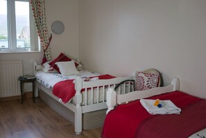 Ein Schlafzimmer mit einem weißen Bett, roter Bettwäsche, einem kleinen Nachttisch, einem Heizkörper und einem Fenster mit Vorhängen.