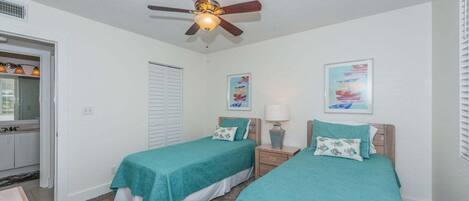 Une chambre avec deux lits simples, un ventilateur de plafond, une porte avec une fenêtre et un mur orné de deux tableaux encadrés.