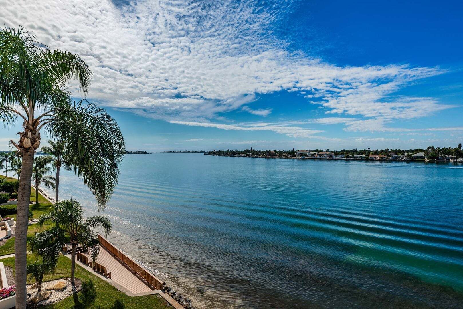A waterfront property with a clear view of the water, a paved walkway, and a row of palm trees.