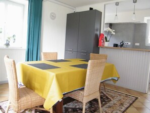 A dining area with a yellow table, four wicker chairs, a grey cabinet, and a kitchen counter with a red bag and a plant.