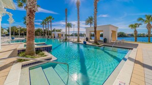 A clear blue swimming pool surrounded by palm trees and a white building with a view of the water. - "Sea La Vie" | Gulf & Lakefront Views | Community Heated Pool | Tennis, Pickleball, Basketball (Panama City Beach)