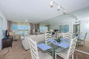A modern hotel room with a glass dining table, white chairs, a sofa, a TV, and a balcony with a view.