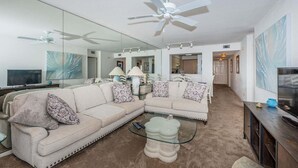 A modern living room with a sectional sofa, a glass coffee table, a ceiling fan, and a large mirror. - Waterfront 2BR condo in St. Petersburg with balcony, pool, and water views. (St Petersburg)