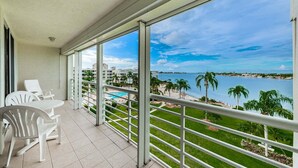 A balcony with a view of a pool and beach, white chairs, and a clear sky. - Waterfront 2BR condo in St. Petersburg with balcony, pool, and water views. (St Petersburg)