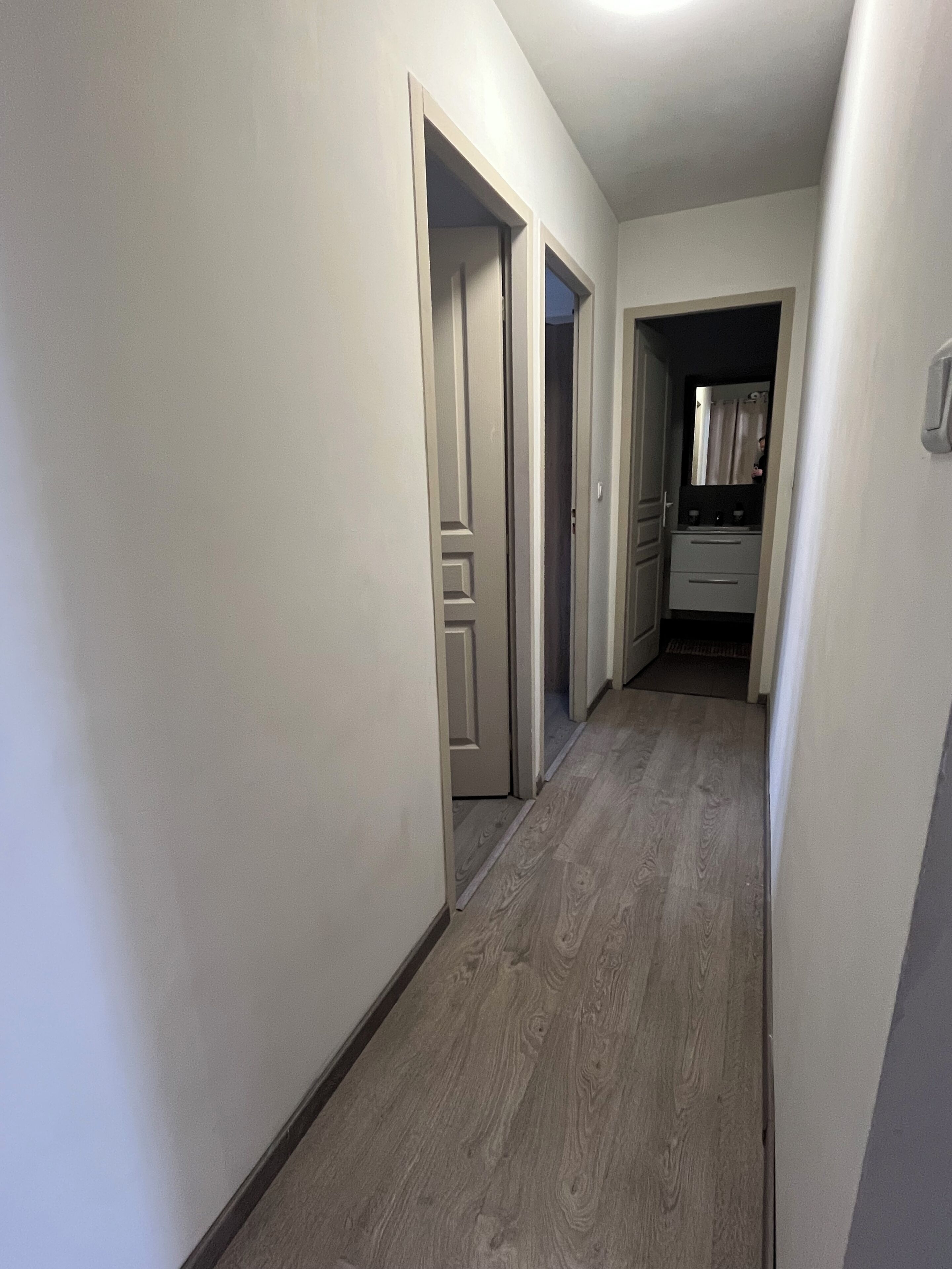 Een smalle gang met houten vloer, witte muren en deuren naar een badkamer en een keuken.