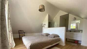 A bedroom with a bed, wooden floor, a chair, a hanging light fixture, a mirror, and a small red trash bin.