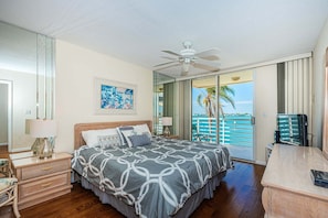 A hotel room with a bed, a desk, a chair, a ceiling fan, a large window with a view of the sea, and a door leading to the balcony.