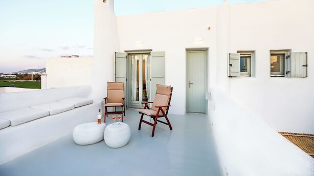 En tagterrasse med en hvid væg, to træstole, en hvid sofa og et lille rundt bord med glas.