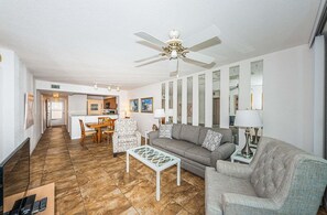 A living room with a sofa, armchairs, a coffee table, and a ceiling fan. There is a dining area with a table and chairs in the background.