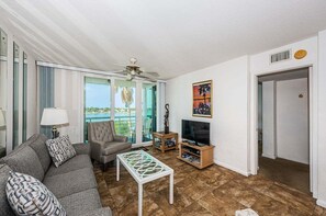 A living room with a sofa, armchair, coffee table, TV stand, and a balcony with a view.