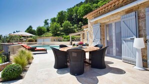 Eine Terrasse mit rundem Tisch und Stühlen, ein Schwimmbad und ein Steinhaus mit großen Garagentoren.