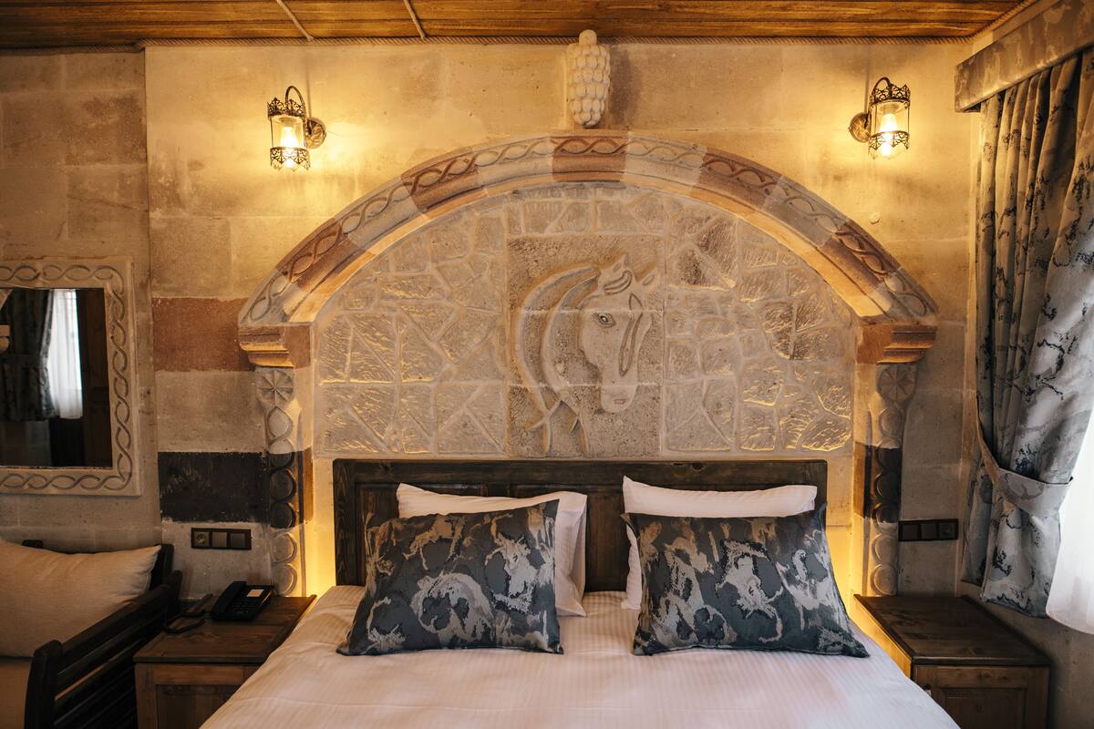 A bedroom with a stone arched headboard, two bedside tables, a bed with patterned pillows, and wall-mounted sconces.