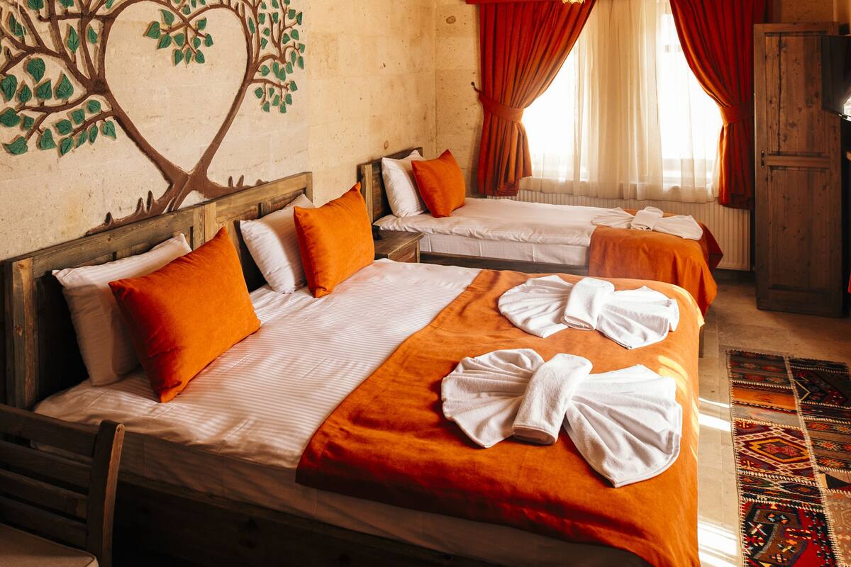 A hotel room with a wooden ceiling, two beds with orange bedding, a heart-shaped wall mural, and a window with red curtains.