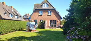 A large brick house with a sloped roof, a small front porch with a table and chairs, and a well-maintained garden with a hedge and flowers.