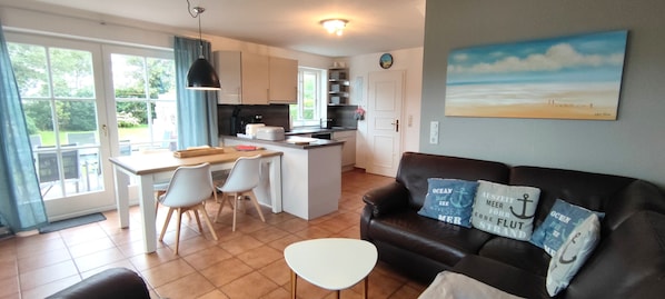 A modern kitchen with a dining area, a living room with a sofa and cushions, and a painting on the wall.
