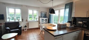 A modern kitchen with a dining area, large windows, and a view of greenery.