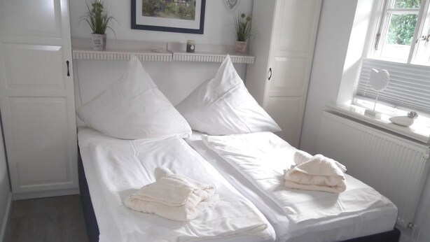 A neatly made double bed with white linens and pillows, a bedside table with a lamp, and a window with blinds. - Rungholtstr. 3 a, apt. jetsam (Wyk auf Föhr)