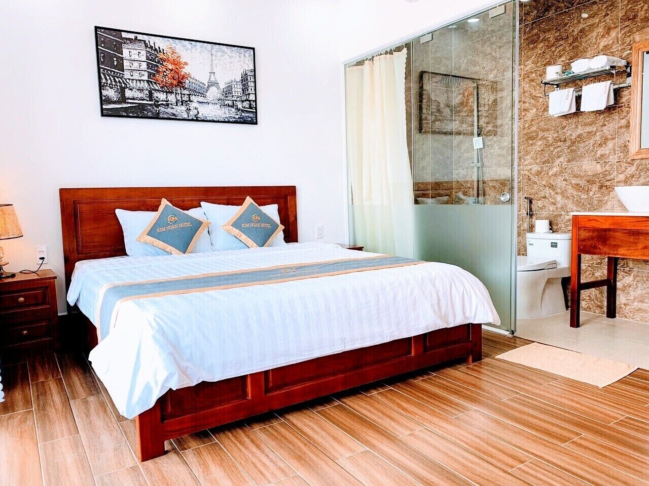 Ein Hotelzimmer mit einem hölzernen Bett, einem Nachttisch, einem Badezimmer mit Dusche und einem an der Wand befestigten Regal mit Handtüchern.