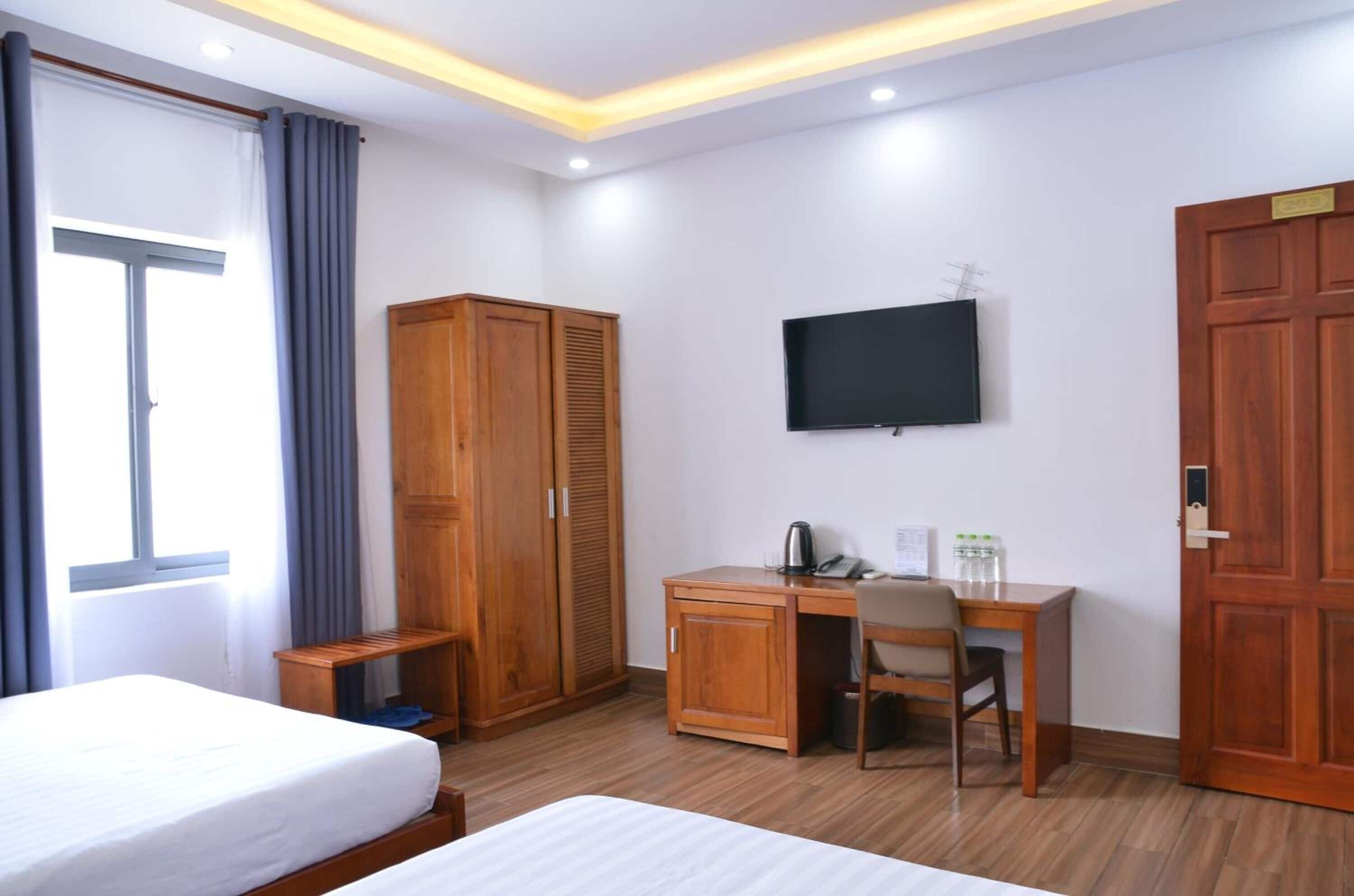 Quarto de hotel com cama, escrivaninha e cadeira, guarda-roupa, televisão e porta.