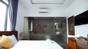 Habitación de hotel con cama, mesita de noche, l ámpara, televisor, escritorio y zona de ducha.